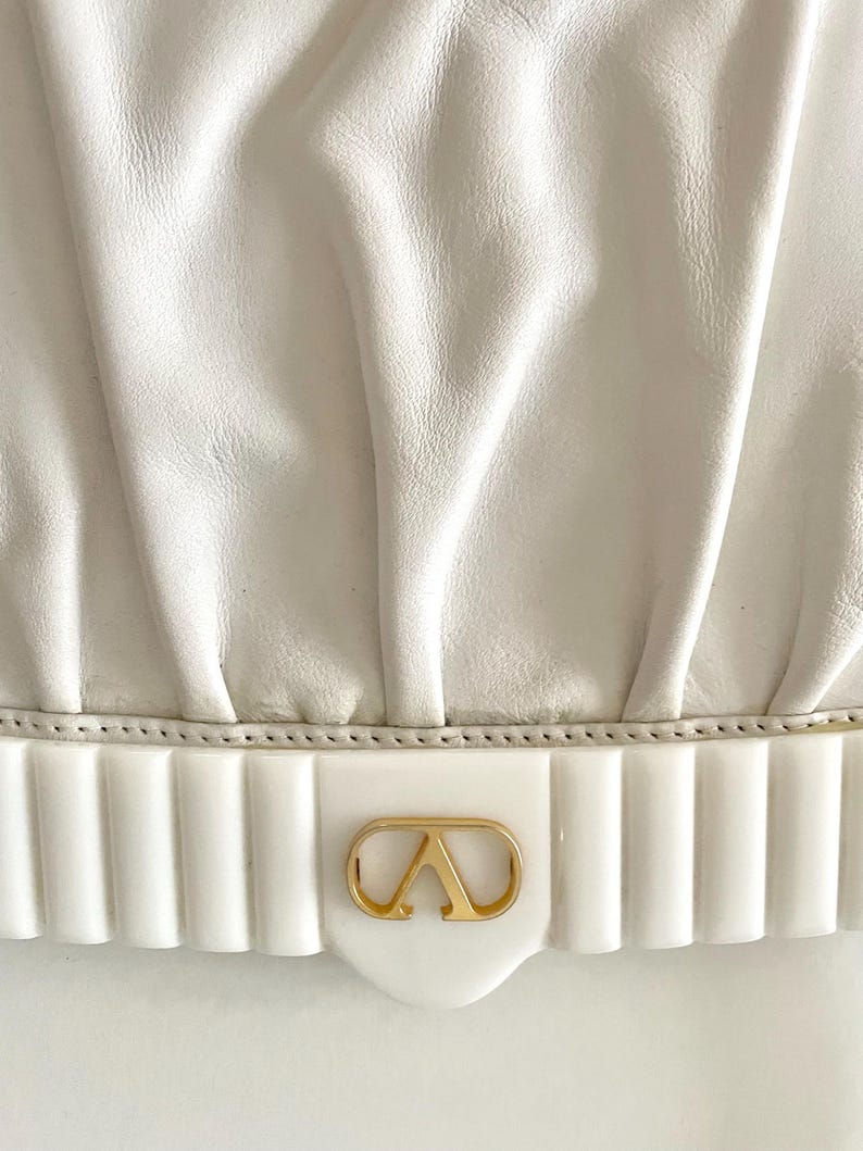 Pode incluir: Bolsa de m&atilde;o de couro branco com um logotipo dourado e uma guarni&ccedil;&atilde;o branca canelada.