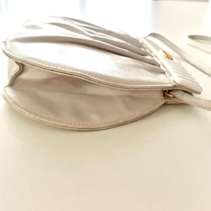 Pode incluir: Uma bolsa de m&atilde;o de cetim branco com um design plissado e um fecho dourado. A bolsa tem uma al&ccedil;a longa e est&aacute; fechada.