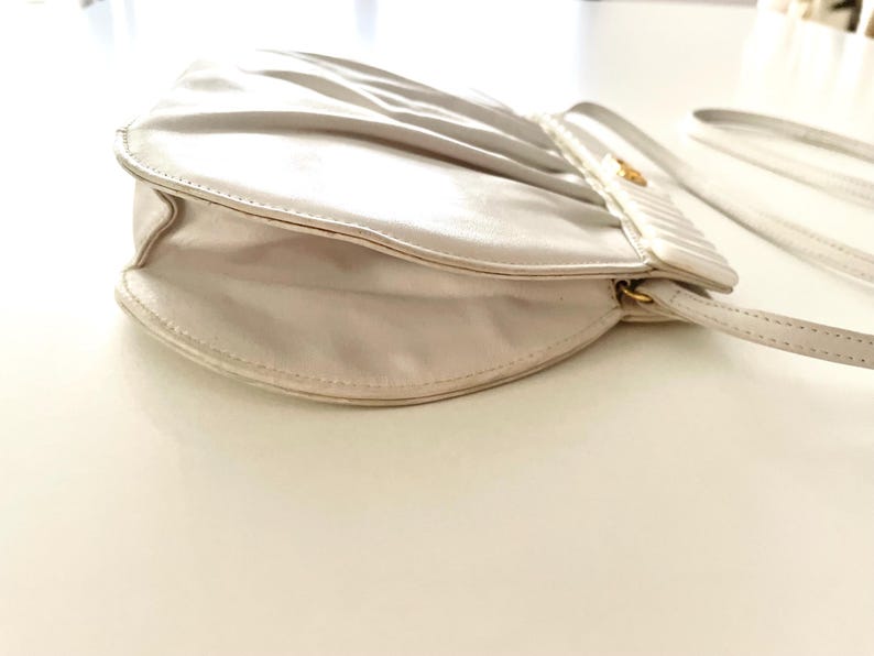 Pode incluir: Uma bolsa de m&atilde;o de cetim branco com um design plissado e um fecho dourado. A bolsa tem uma al&ccedil;a longa e est&aacute; fechada.