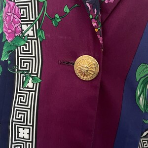 May include: A close-up of a purple blazer with a gold button. The blazer features a floral and geometric pattern in shades of purple, blue, green, and white. The gold button has a sunburst design.