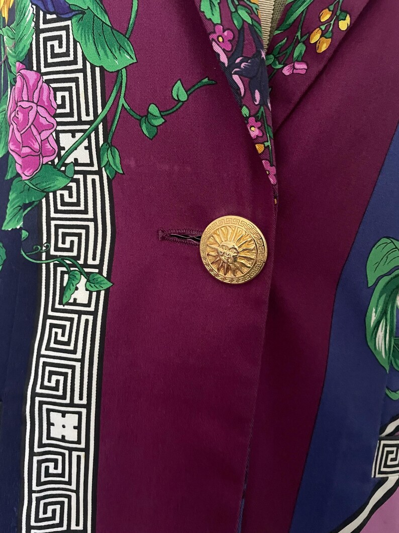 May include: A close-up of a purple blazer with a gold button. The blazer features a floral and geometric pattern in shades of purple, blue, green, and white. The gold button has a sunburst design.
