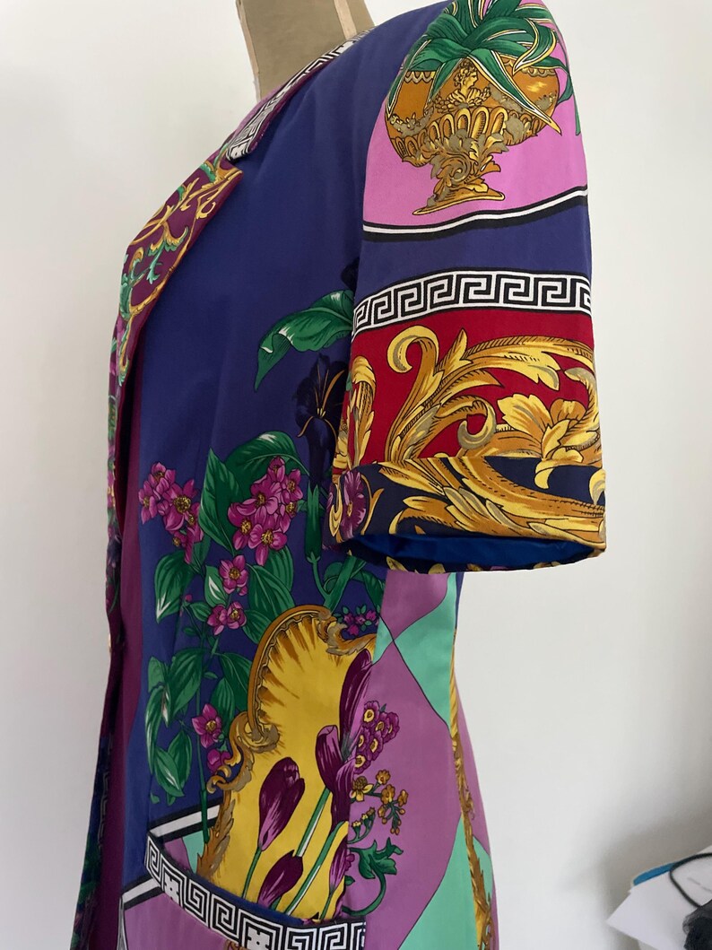 May include: A vintage, knee-length dress in shades of blue, purple, and yellow. The dress features a vibrant floral and ornamental pattern, with gold accents and a Greek key border. The sleeves are short, and the dress has a tailored fit.
