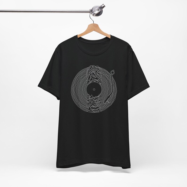 May include: A black short-sleeve t-shirt featuring a white line art graphic on the chest. The design depicts a vinyl record with a tonearm, where the grooves of the record form a mountain-like wave pattern. The shirt is displayed on a wooden hanger.