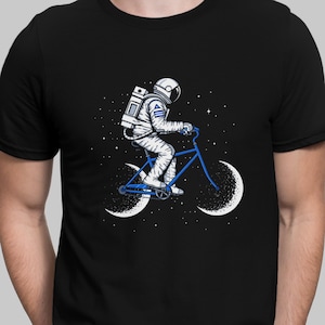 Könnte beinhalten: Ein schwarzes T-Shirt mit einem Motiv eines Astronauten, der ein Fahrrad mit mondförmigen Rädern in einem sternenklaren Himmel fährt.