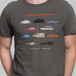 Op de afbeelding: Een antracietgrijs t-shirt met een pixel art afbeelding van diverse iconische voertuigen. De tekst bovenaan luidt RETRO RIDEZ en onderaan staat Press Start! Credits:02. Het ontwerp bevat onder meer een witte kever, een zwarte bus en een rode auto.