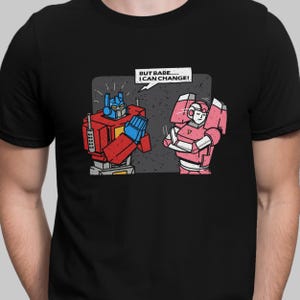 May include: Black t-shirt featuring a cartoon illustration of two robots. One robot is red, blue, and gray, with its hand raised. The other robot is pink. Text above the robots reads, "BUT BABE..... I CAN CHANGE!"