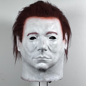 May include: A white mask with a realistic face, brown hair, and a stand. The mask has a neutral expression with closed eyes.