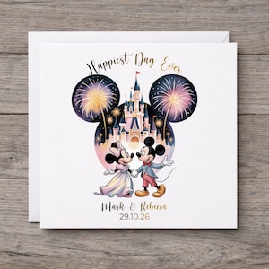 Puede incluir: Una tarjeta de felicitación cuadrada con una ilustración de dos personajes de ratón tomados de la mano frente a un castillo con fuegos artificiales. El texto dice Happiest Day Ever, Mark & Rebecca, y la fecha 29.10.26 en letras doradas.