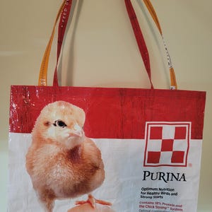 May include: A tote bag made from a red and white Purina feed bag. The bag features a large image of a chick standing on an egg. The bag has orange and red straps with the words "FEED GREATNESS" printed on them.