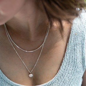 Könnte beinhalten: Eine silberne Kettenkette mit einem kleinen, runden Anhänger. Die Kette ist über eine zweite, dünnere Kette gelegt.