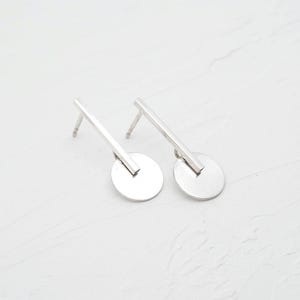 Può includere: Un paio di orecchini d'argento con un design geometrico semplice. Ogni orecchino presenta un piccolo disco rotondo con una linea retta che si estende da esso.