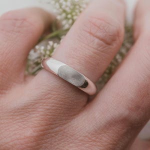 Könnte beinhalten: Ein silberner Ring mit einem Fingerabdruck-Design. Der Ring wird an einem Finger getragen, mit einem hellen Hintergrund. Der Ring hat eine glatte, polierte Oberfläche und eine abgerundete Form. Der Fingerabdruck ist eine graue ovale Form.