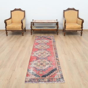 May include: A vintage, distressed runner rug with a red and faded color palette. The rug features a central pattern of geometric shapes in shades of beige, blue, and black. Two antique armchairs and a small table are in the background.