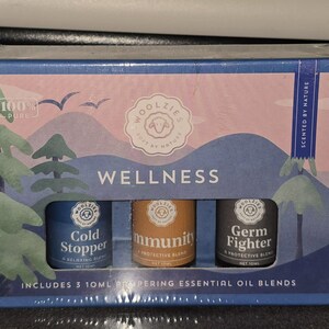 Puede incluir: Un juego de tres mezclas de aceites esenciales de 10 ml. La caja es azul y rosa con un diseño temático de la naturaleza. Las botellas están etiquetadas como "Cold Stopper", "Immunity" y "Germ Fighter". El texto "Wellness" se muestra en la caja.