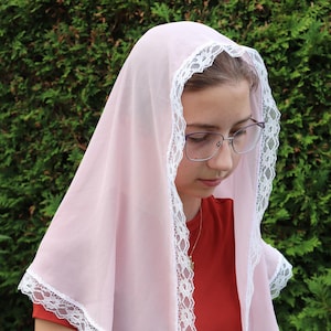 May include: A light pink head covering made of sheer fabric with a white floral lace trim along the edges. The triangular-shaped veil drapes over the head, featuring a delicate scalloped lace border that frames the face and extends down the sides.