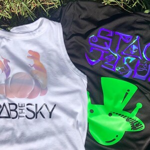 Puede incluir: Dos camisetas sin mangas blancas con diseños gráficos coloridos. La camiseta de la izquierda presenta un dinosaurio de color arcoíris y una criatura alada con el texto "DAB THE SKY". La camiseta de la derecha presenta un extraterrestre verde con un sombrero de copa y el texto "SPACE JESUS" en morado.
