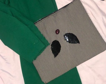 Puede incluir: Un blazer verde con un clutch estampado en blanco y negro. El clutch tiene una cremallera negra y está decorado con adornos negros en forma de hoja y una pequeña joya redonda roja. Se ve un artículo blanco y crema.