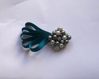Puede incluir: Broche decorativo con un racimo de perlas de imitación en tonos grises y blancos, acentuado con un brillante central. El broche está unido a un abanico de cinta de raso verde azulado.