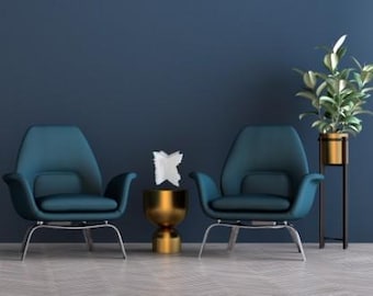 Puede incluir: Dos sillones verde azulado con patas plateadas se enfrentan, flanqueando una pequeña mesa auxiliar dorada con un objeto decorativo blanco. Una planta en maceta en una maceta dorada se encuentra en un soporte negro a la derecha, todo contra una pared azul oscuro.