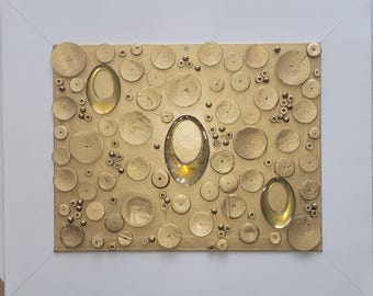 Puede incluir: Obra de arte abstracta con una superficie texturizada dorada, que incorpora elementos circulares de varios tamaños y tres acentos ovalados dorados transparentes. La obra está enmarcada en un marco rectangular blanco.