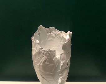 Puede incluir: Una escultura abstracta blanca con una apariencia texturizada y arrugada. La escultura está montada sobre una base cuadrada negra. El fondo es una pared verde oscuro y la escultura está sobre una superficie blanca.