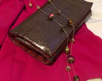 Puede incluir: Un clutch marrón texturizado con un collar de cadena dorada con cuentas de colores. El collar está drapeado sobre el clutch y un blazer rosa brillante. El blazer está doblado y tiene un forro blanco.