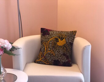 Puede incluir: Cojín decorativo cuadrado con un estampado floral en tonos verde oliva, amarillo y burdeos. El cojín está colocado sobre un sillón blanco, con una pared rosa de fondo. El diseño del cojín presenta una flor grande y estilizada.