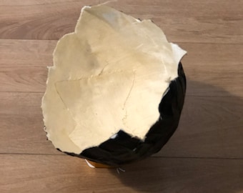 Puede incluir: Un objeto esférico parcialmente construido, con un exterior negro y un interior beige claro. El objeto parece estar hecho de materiales en capas, con una textura rasgada, similar al papel, en el interior. Descansa sobre una superficie de madera.
