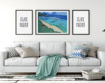 Puede incluir: Un salón con un sofá blanco, cojines y una manta turquesa. Encima del sofá hay tres obras de arte enmarcadas, incluyendo una pintura de paisaje colorida. Una pequeña planta y una lámpara completan la decoración.