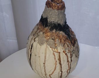 Puede incluir: Un jarrón artesanal texturizado con un diseño único. El jarrón presenta una base blanca con goteos marrones, una banda negra y una parte superior texturizada gris y dorada. El jarrón es un elemento de decoración artística.