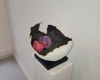 Puede incluir: Escultura abstracta en forma de huevo agrietado. El exterior es blanco con una superficie texturizada, el interior es negro con ribete dorado. Dos flores pintadas en rosa y morado están dentro. La escultura descansa sobre una base negra.