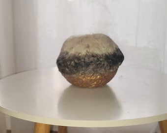 Puede incluir: Un jarrón decorativo esférico con una superficie texturizada. El jarrón presenta un degradado de colores, pasando del dorado en la base al negro y luego al beige en la parte superior. El jarrón está sobre una mesa blanca.