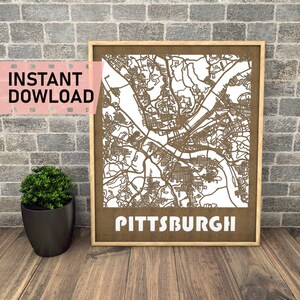 May include: A brown wooden frame with a white background. The frame contains a detailed laser cut design of the city of Pittsburgh. The city name is written in white letters at the bottom of the frame.