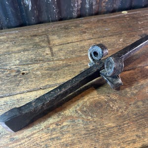 May include: A hand-forged black iron metal spike featuring a pointed tip and a decorative scroll detail near the center. The object has a textured, dark finish with a flat, widened base and a tapered, sharp end, showcasing a rugged, industrial appearance.