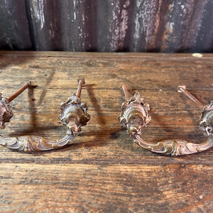 Op de afbeelding: Twee antieke bronzen ladehandgrepen met sierlijke bloemmotieven. Elke handgreep heeft een gebogen handvat en decoratieve uiteinden. De handgrepen staan op een rustiek houten oppervlak, wat hun vintage uitstraling benadrukt.