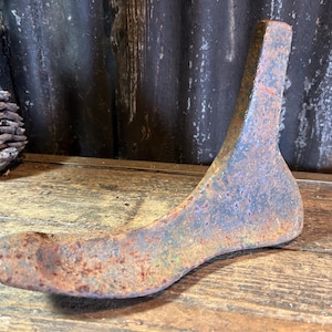 May include: A vintage, rusty metal shoe last, showing signs of age and wear. The shoe form is a foot-shaped mould used in shoemaking. The object is resting on a wooden surface, with a pine cone in the background.