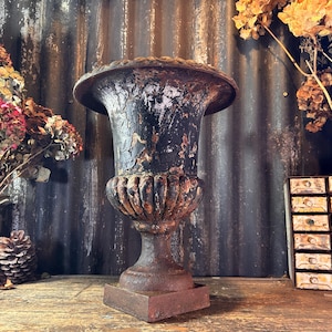 Puede incluir: Una urna oscura y desgastada, con una parte superior ancha y acanalada y una base de pedestal. La urna tiene un acabado envejecido, lo que sugiere edad y uso en exteriores. El jarrón decorativo es probablemente de hierro fundido.