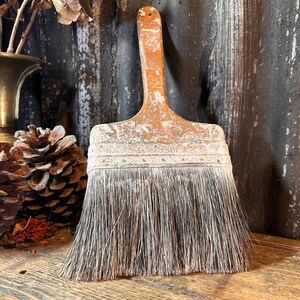 Può includere: Un pennello invecchiato con manico in legno e setole usurate. Il pennello è di colore marrone caldo, che mostra segni di utilizzo. Le setole sono un misto di grigio e bianco. Il pennello viene probabilmente utilizzato per dipingere o per progetti artistici.