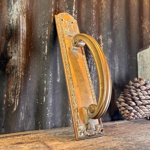 Pode incluir: Uma maçaneta de porta antiga de latão com uma placa traseira retangular e uma alça curva. A maçaneta tem detalhes ornamentados e uma pátina quente e envelhecida. A maçaneta está encostada em um fundo escuro e desgastado.