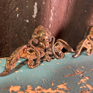 May include: Two antique, ornate, gold-toned metal angel wings or decorative embellishments. The detailed castings feature cherubic faces and stylized wings. The background is a distressed, weathered surface with shades of brown and teal.