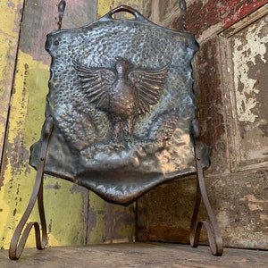 May include: A vintage metal fire screen with a hammered finish and a raised design of a bird with outstretched wings. The screen is mounted on a simple black metal stand.