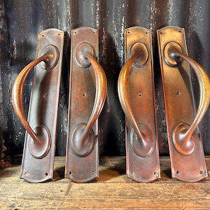 May include: Four antique bronze door handles with curved handles and rectangular backplates. The handles have a warm, reddish-brown patina, suggesting age and use. The hardware is likely for a vintage or rustic door.