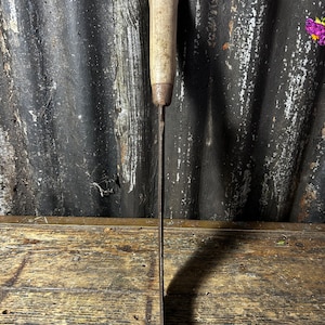 May include: A long, thin, rusty metal blade with a wooden handle. The blade is straight and has a slight curve at the end. The handle is brown and has a smooth surface.