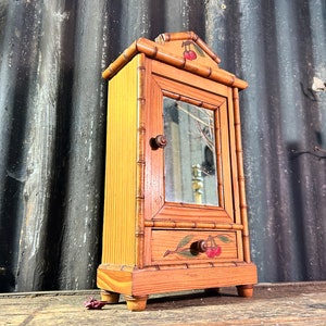 Può includere: Un piccolo mobiletto in legno con una porta a specchio e un cassetto. Il mobiletto è dipinto con una tinta marrone chiaro e presenta un motivo a ciliegie sulla parte superiore e sul cassetto.
