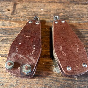 May include: Two vintage brown leather pulleys with metal hardware. Each pulley has a loop at the top and is marked "MADE IN ENGLAND" and "EASWORTH" or "LEWIN M". The pulleys are set against a weathered wooden surface.