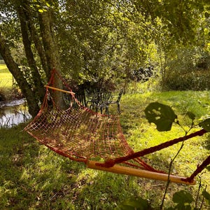 May include: A red hammock hangs between two trees over a small stream. The hammock is made of a woven net material and has a wooden frame. The hammock is surrounded by green grass and trees.