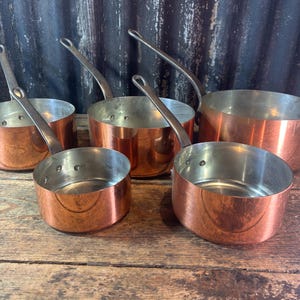 Pode incluir: Um conjunto de quatro panelas redondas de cobre com interiores de metal prateado e cabos longos de metal rebitados. As panelas variam em tamanho e estão dispostas em grupo, cada uma com um exterior de cobre polido.