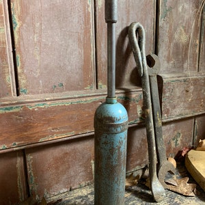 May include: A vintage blue and rusty metal grease gun with a long metal handle. The grease gun is sitting on a wooden surface with other tools.