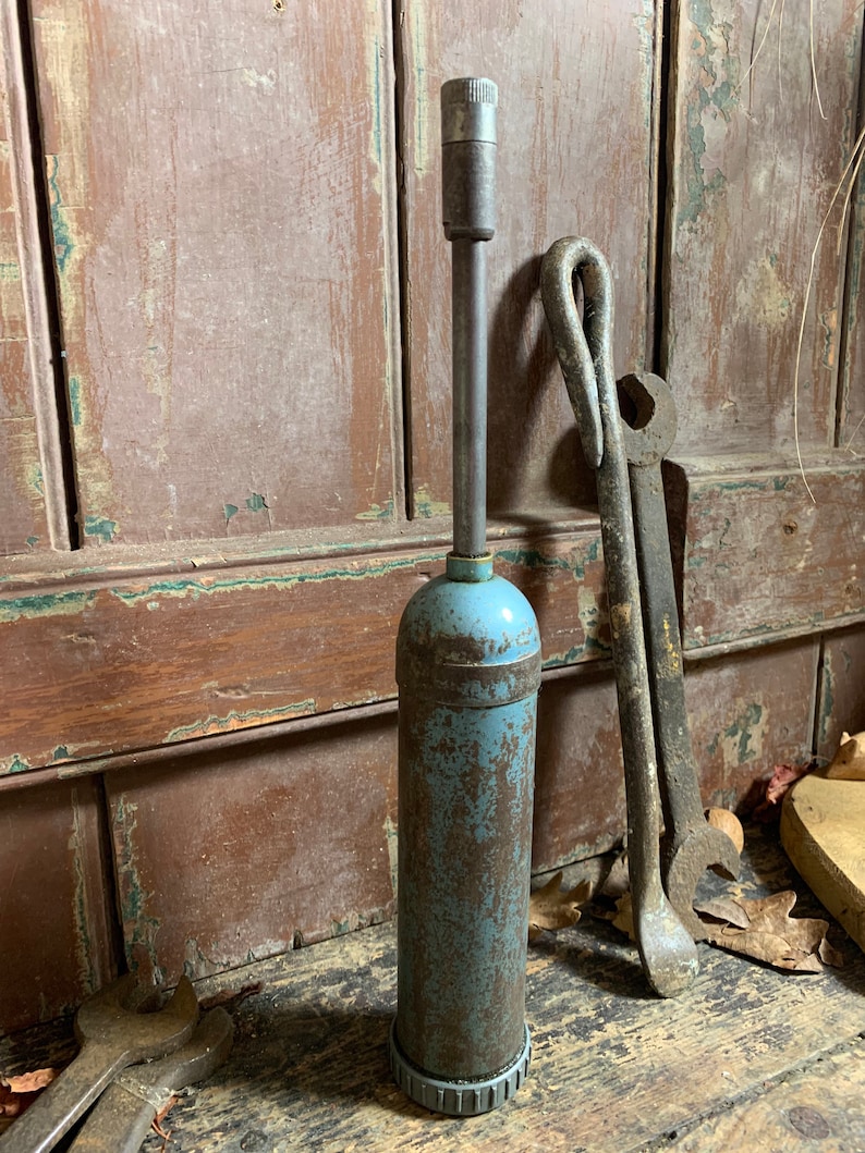 May include: A vintage blue and rusty metal grease gun with a long metal handle. The grease gun is sitting on a wooden surface with other tools.