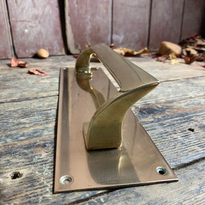 May include: A polished brass door handle with a long, rectangular backplate. The handle has a unique, curved design.